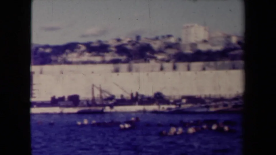 Final 1945 France frame showing boats on blue water below a pale waterfront wall, cranes, and hillside buildings in soft 8mm color.