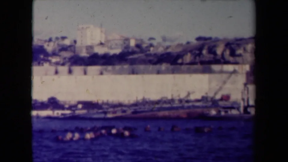 Distant 1945 French port view with a docked cargo ship, cranes, white quay wall, and hillside buildings seen through soft film grain.
