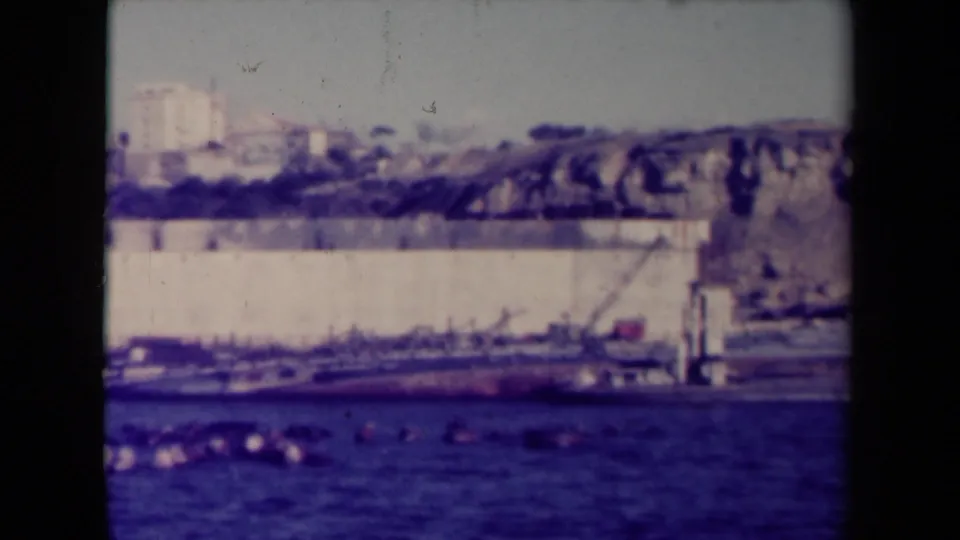 1945 France harbor frame showing cargo vessels near a pale quay wall, cranes, hillside structures, and blue water in the foreground.