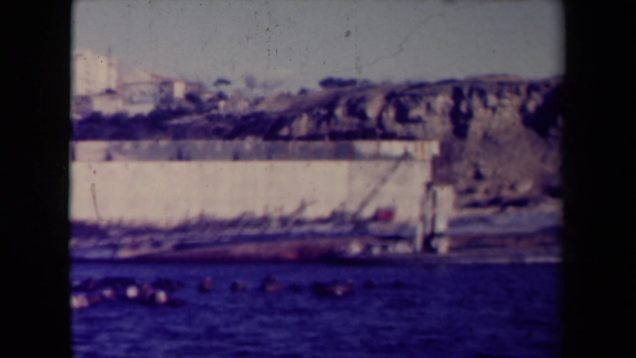 Large vessel lying on its side near a shoreline wall with water in the foreground.