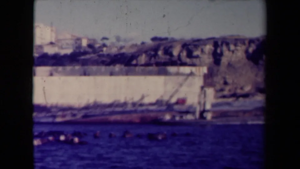 1945 France 8mm frame showing a large capsized vessel beside a coastal wall, with dark water and distant buildings visible.
