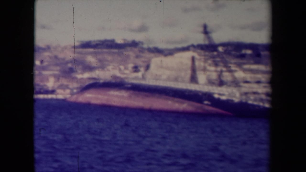 Capsized ship hull partly submerged beside a coastal wall and distant hillside.