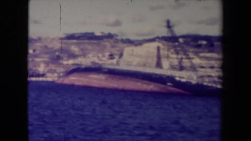 1945 France 8mm frame with the reddish underside of a capsized ship visible against water, shoreline structures, and low hills.