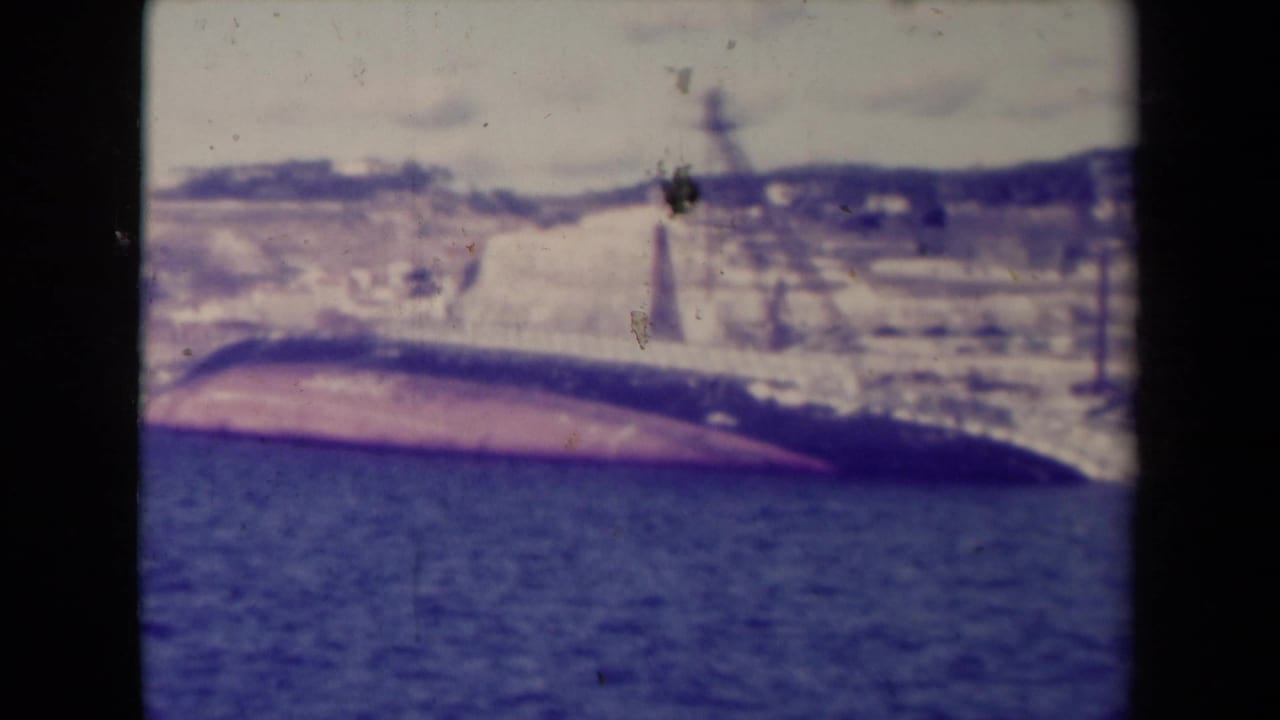 1945 France 8mm frame showing the broad side of a capsized ship aground near the shoreline, with water and hills behind it.