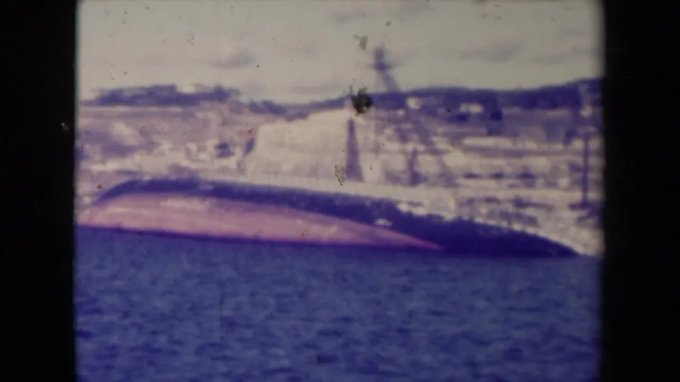 1945 France 8mm frame showing the broad side of a capsized ship aground near the shoreline, with water and hills behind it.