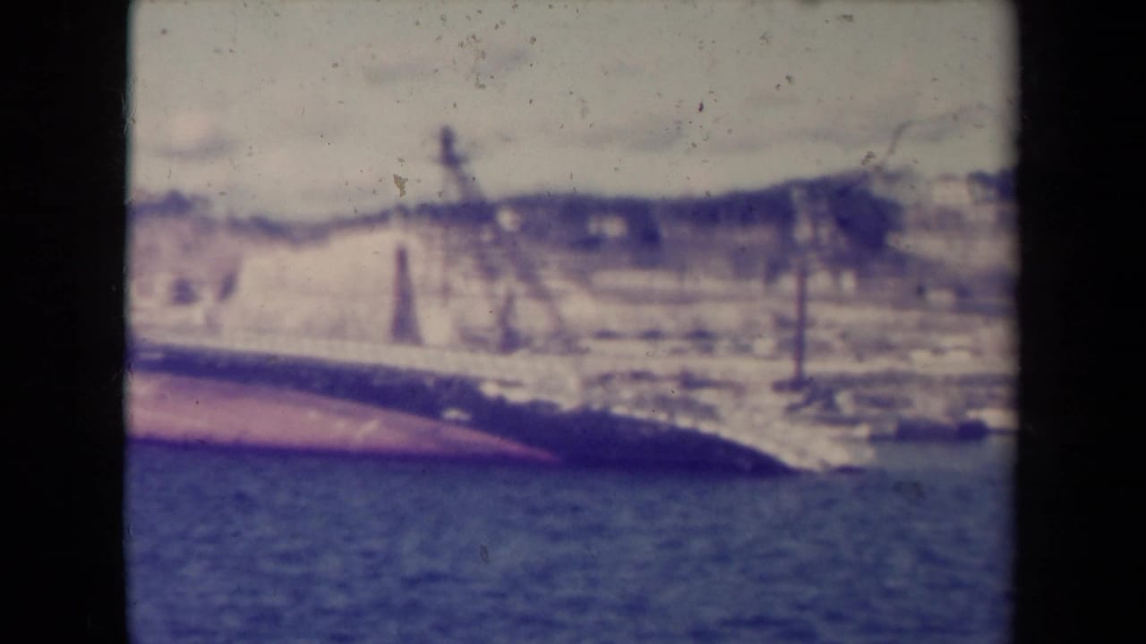 1945 France 8mm frame of a capsized vessel beside a coastal wall, with dark water, low hills, and aged film texture.