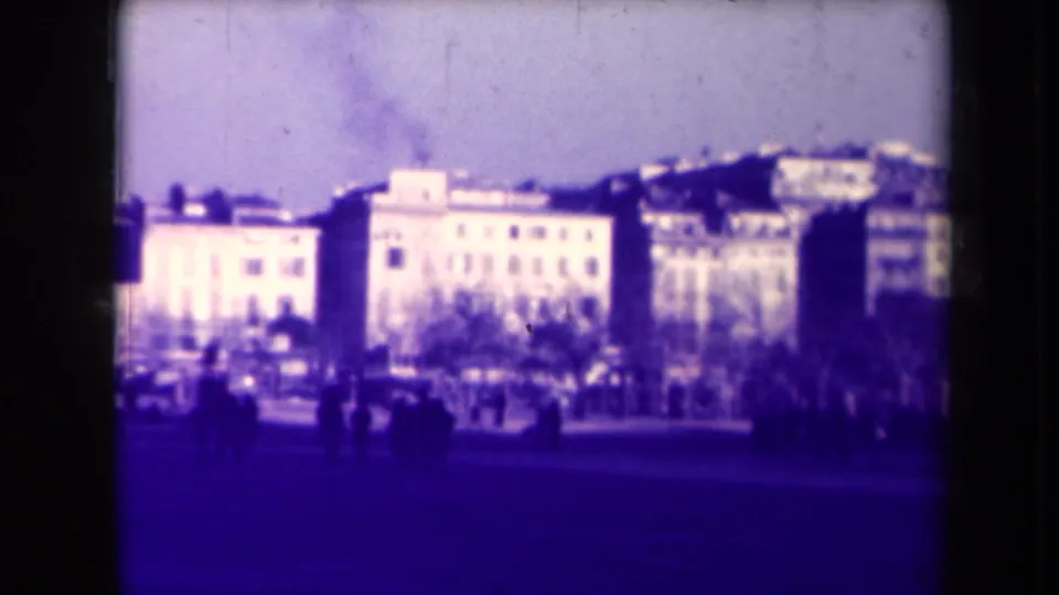 1940s France street scene showing a broad open area, pale multi-story buildings, bare trees, and small distant figures walking or standing.
