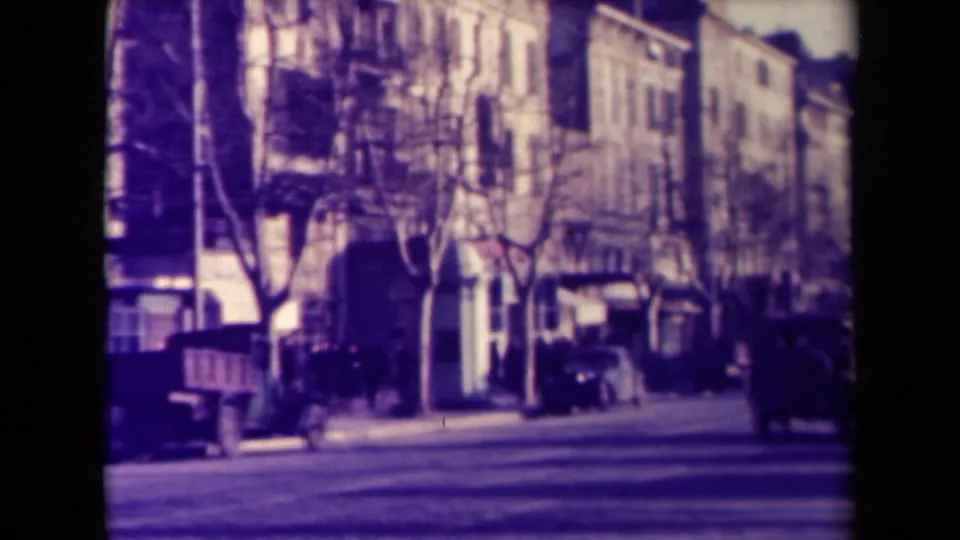 1945 France street view with parked rounded cars, bare trees, pedestrians, shopfronts, and apartment facades.