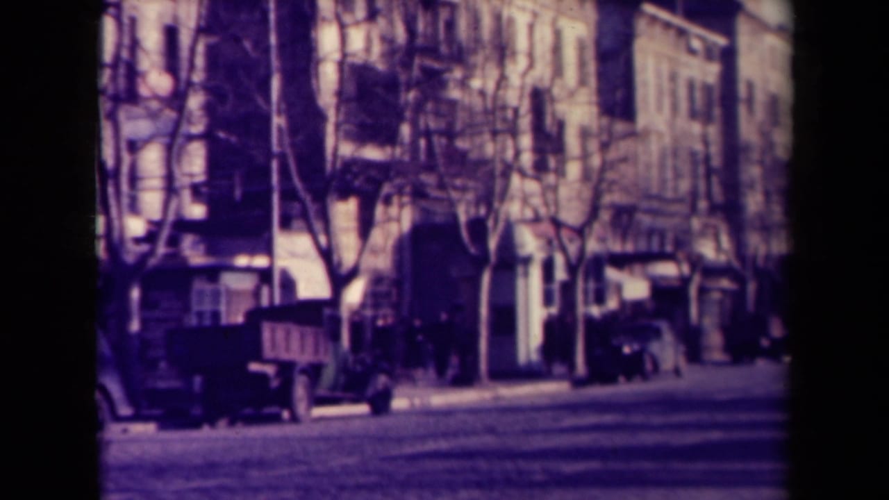 Street corner with classic cars, pedestrians, trees, and tall building facades.