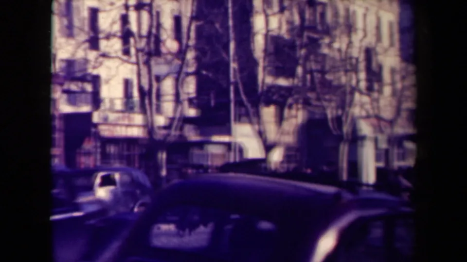 1945 France street frame with a dark car crossing the foreground, a parked rounded car, trees, balconies, and storefronts.