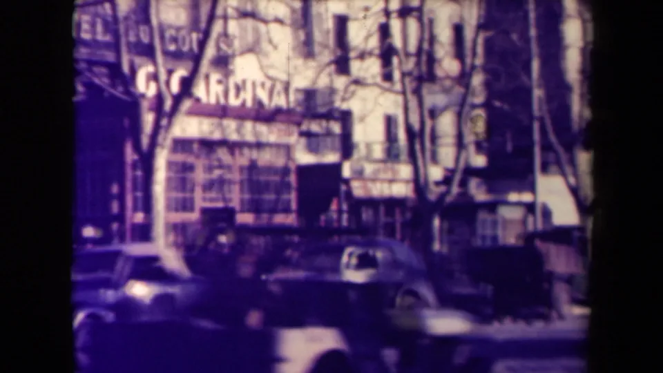 1945 France urban storefront view with a large partially legible sign, parked classic cars, pedestrians, trees, and balconies.