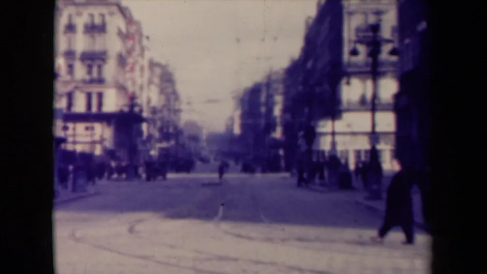 Wide 1940s French urban street view with pedestrians, multi-story buildings, a broad roadway, and soft distant traffic or movement.
