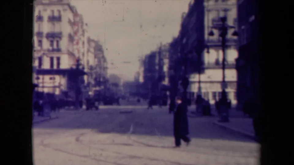 1940s France street scene with a dark-coated pedestrian crossing in the foreground and other people along the sidewalks.