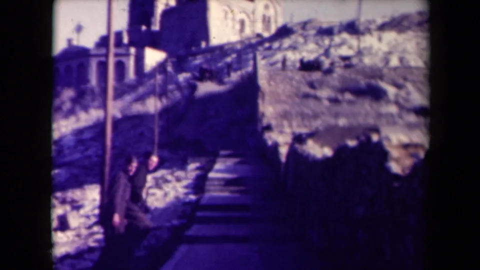 Two people in 1940s France appear to climb a long outdoor staircase toward a towered building on a rocky hillside.
