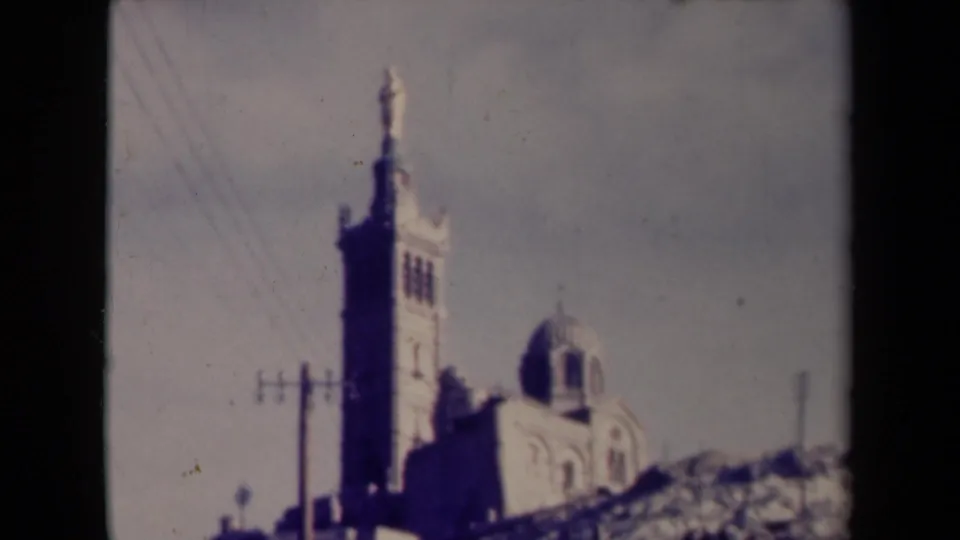 Soft 8mm frame of the hillside church in France, emphasizing the tall tower, dark dome, overhead wires, and pale masonry.