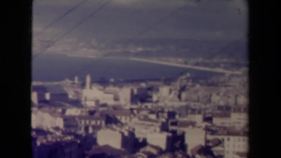 1940s France travel footage widens to an elevated view of town rooftops, a church-like structure, hills, and a curving coastline.