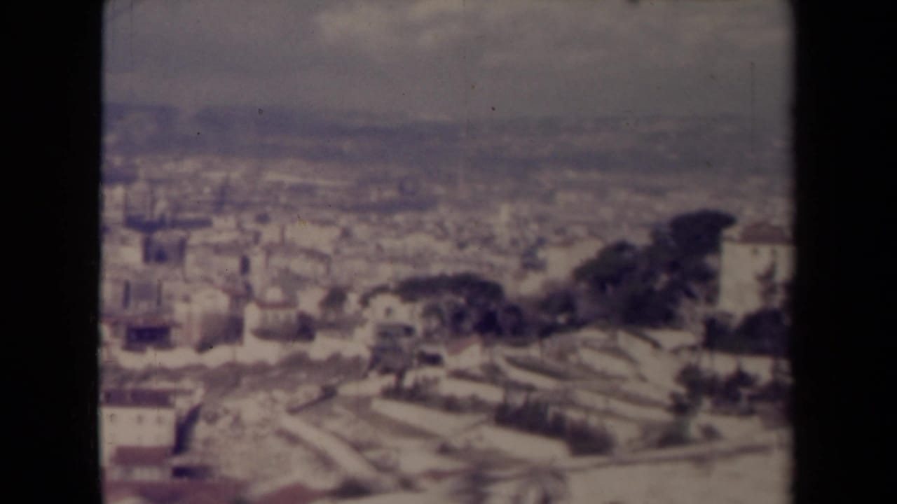 Soft 8mm view over a pale hillside town with clustered buildings, terraces, trees, and distant haze.
