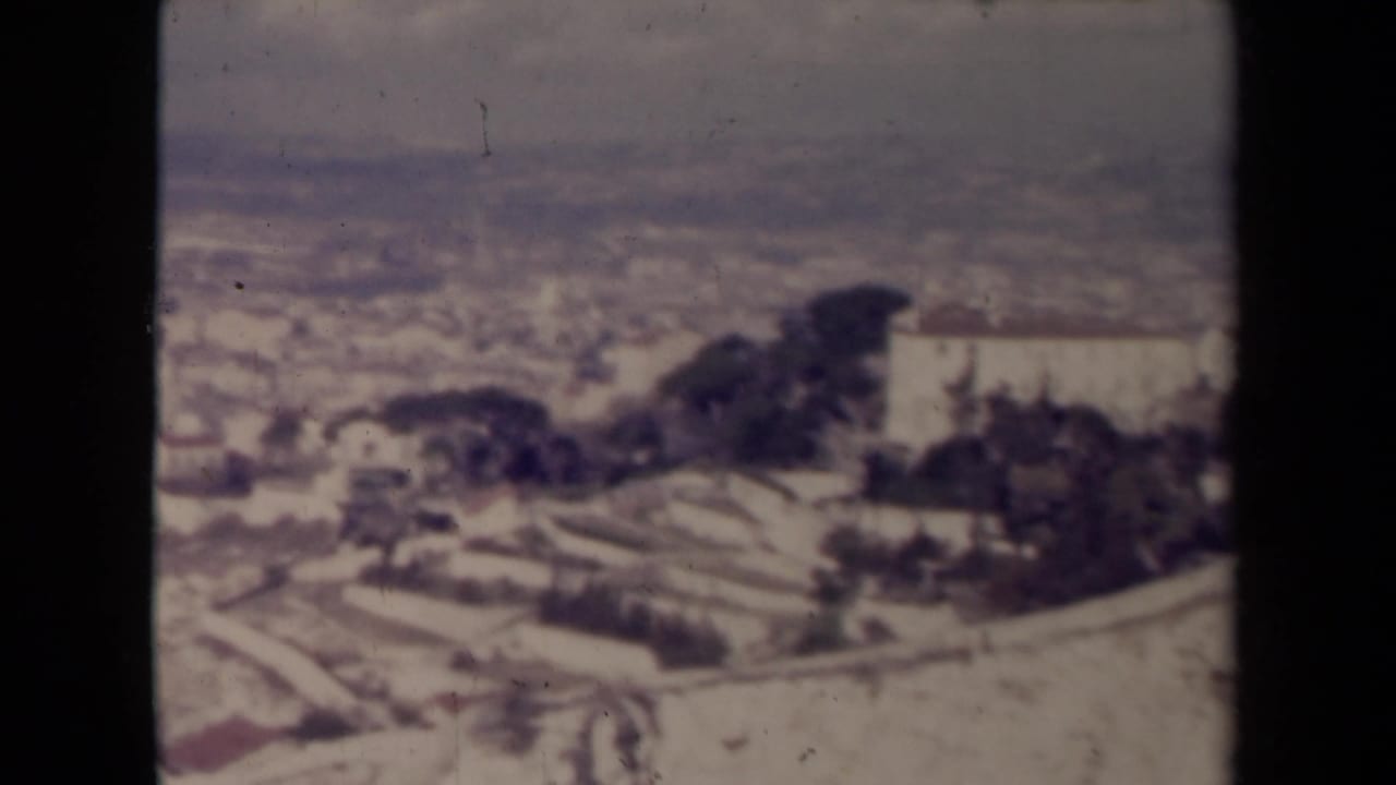 High town panorama with pale buildings, trees, terraces, and a large light-colored structure on the right.
