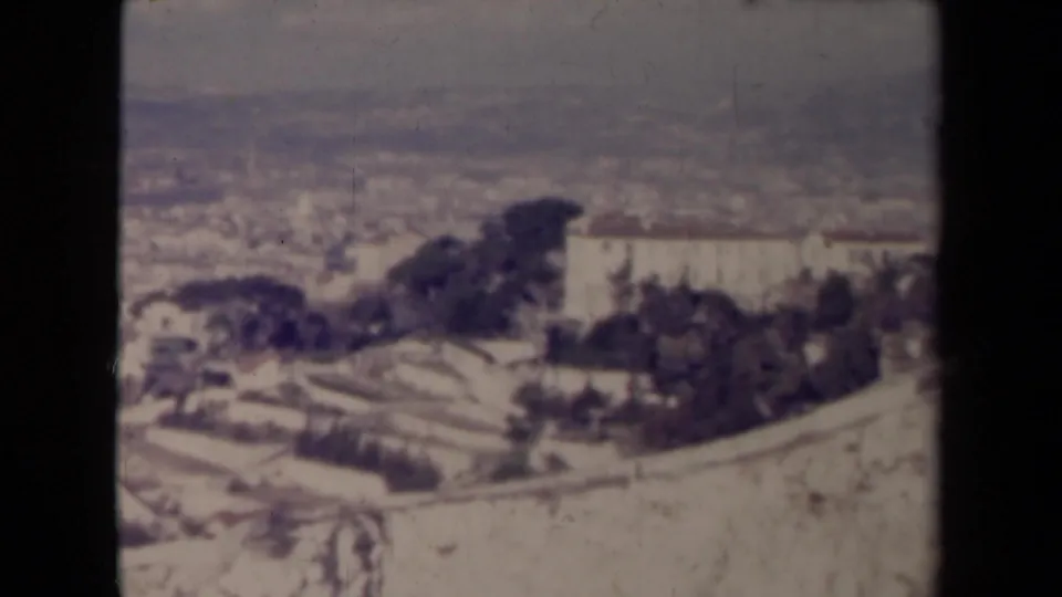 1945 8mm frame from an elevated viewpoint, showing a likely French town with terraced ground, rooftops, dark vegetation, and a pale building mass.