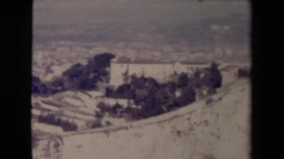 Likely French townscape in a 1945 8mm home movie, with a broad pale hillside foreground, clustered rooftops, dark trees, and distant haze.