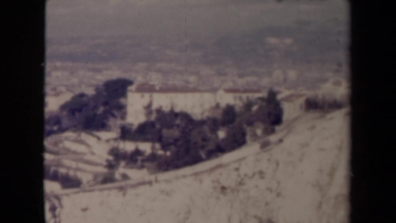 Soft elevated view of a pale building complex on a hillside with trees and distant landscape.