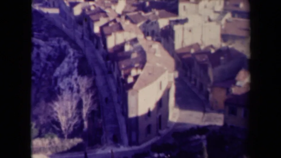 1940s France frame looking down over clustered town buildings, terracotta roofs, a narrow lane, and bare trees.