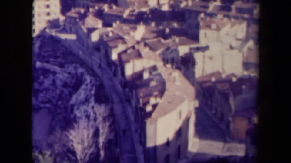 1940s France home-movie frame with a blurred elevated view of rooftops, pale walls, trees, and a larger white building.