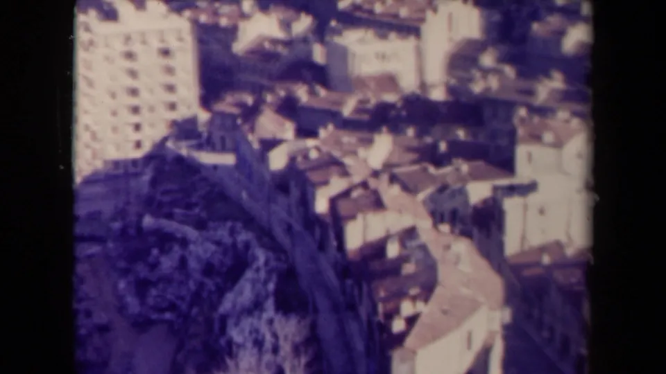 1940s France frame showing dense rooftops and pale town buildings from above, with a taller block along the left edge.
