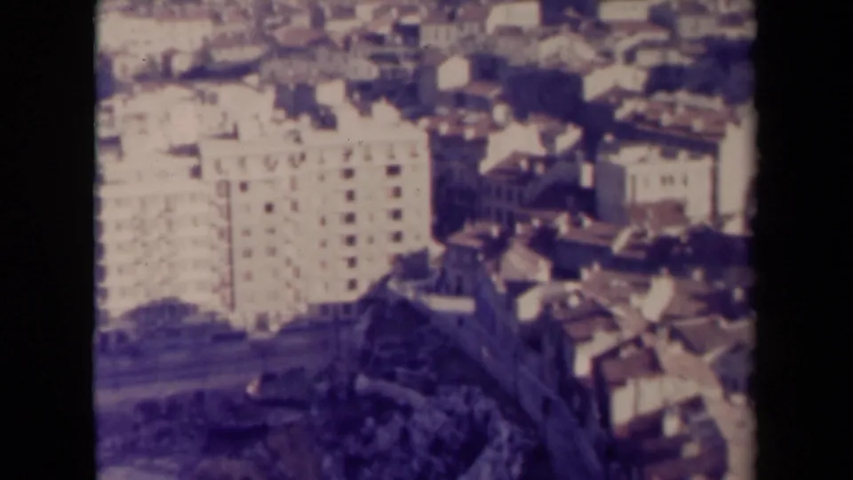 Aged 8mm view over a French urban slope, with a pale building, rooftops, and dark foreground terrain or wall detail.