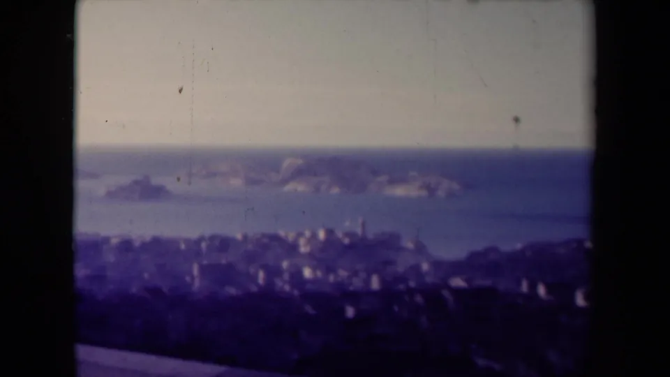 1945 France 8mm frame showing a hazy coastal city below rocky offshore islands, seen through dark film borders.