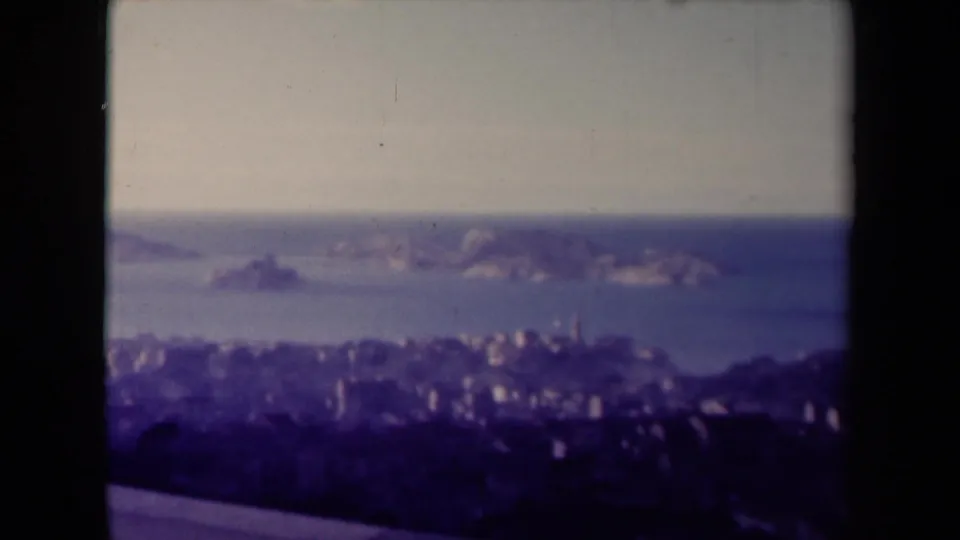 1945 France home-movie frame with a soft distant city, blue sea, and rocky islands under a hazy sky.