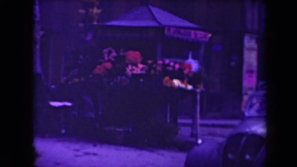 1945 France frame showing a dark street kiosk or market stall with flowers or goods near a snowy sidewalk and parked vehicle edge.