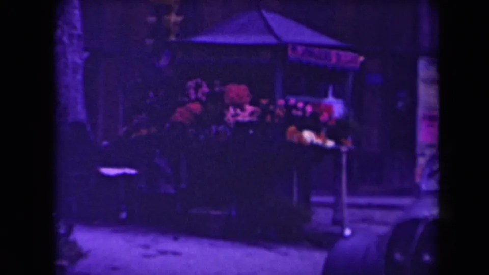 1945 France home-movie frame showing a dark canopied stall with colorful goods near a narrow urban passage.