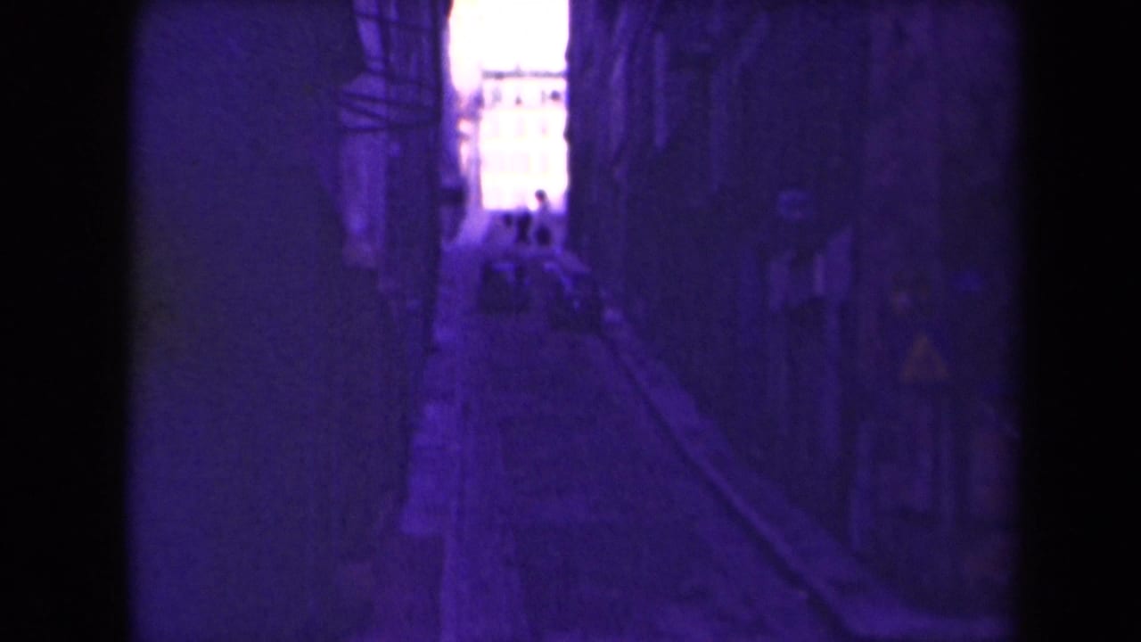 1945 France home-movie view down a narrow alley toward bright daylight with small distant figures visible.