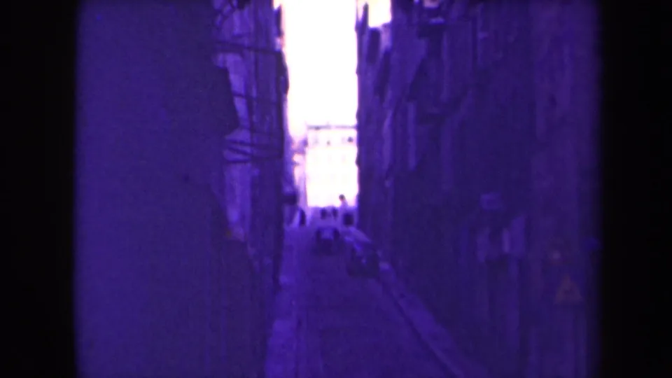 1940s France home-movie frame showing a narrow urban alley in purple-shifted color, with tall walls and a bright street opening beyond.