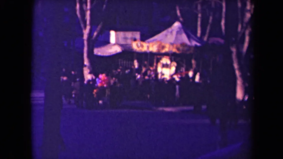 Nighttime fairground view in France, with a glowing carousel canopy, bare trees, and a crowd seen in soft-focus 8mm color.