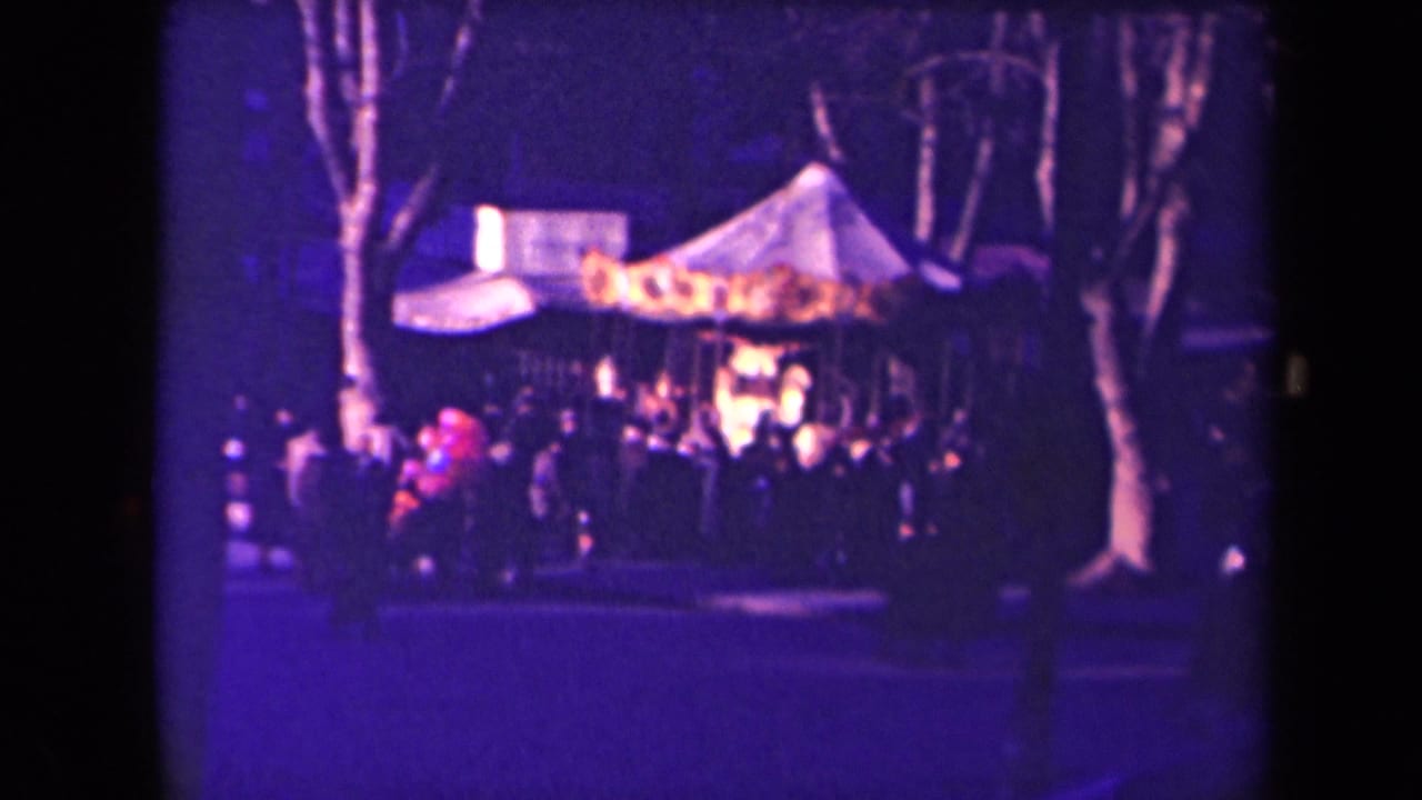 Soft-focus 1940s France fairground frame showing people gathered around a warmly lit carousel beneath tall bare trees.