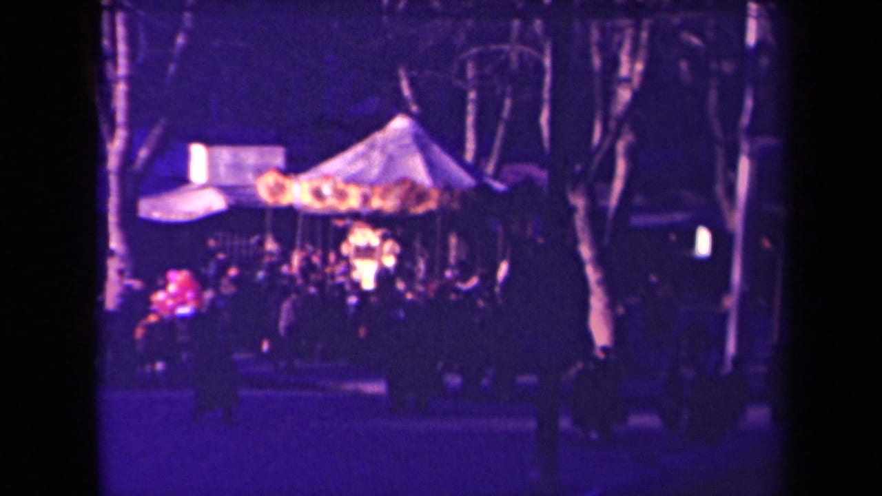 1940s France home-movie frame of a nighttime crowd around a lit carousel, with trees and strong purple color fading across the scene.