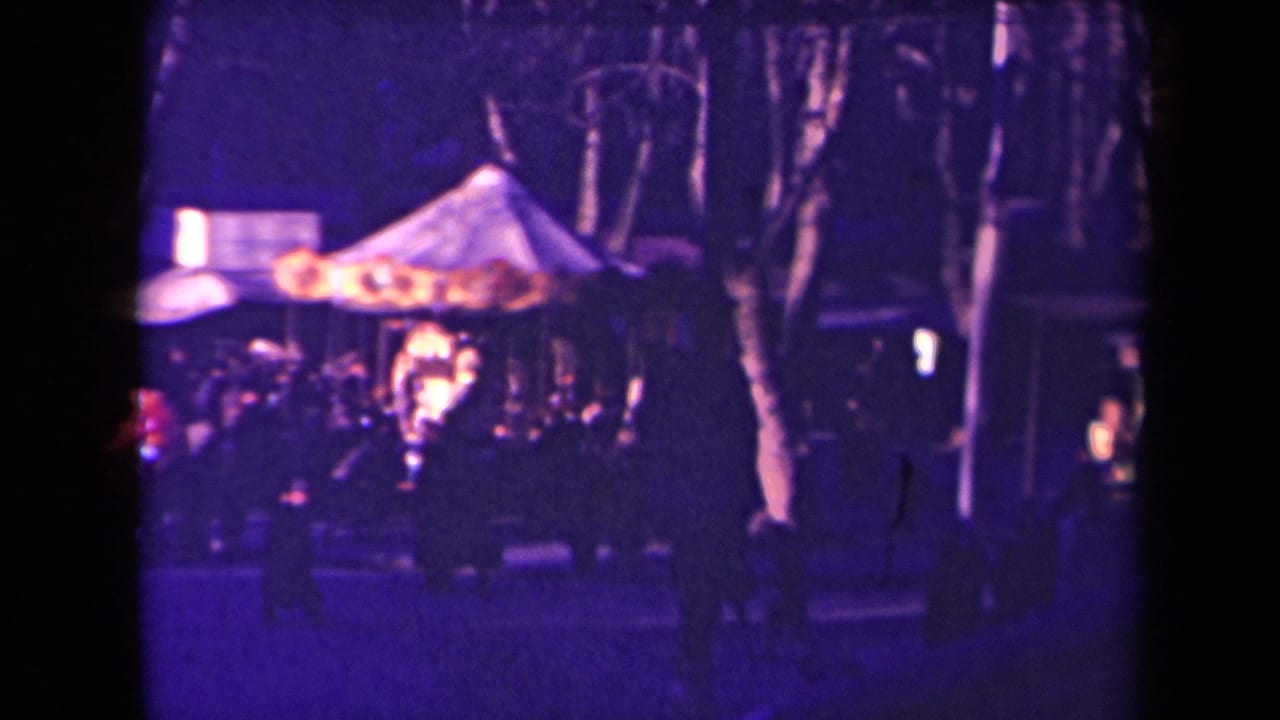 Night carnival scene with a lit carousel tent, silhouetted trees, and people in the foreground.