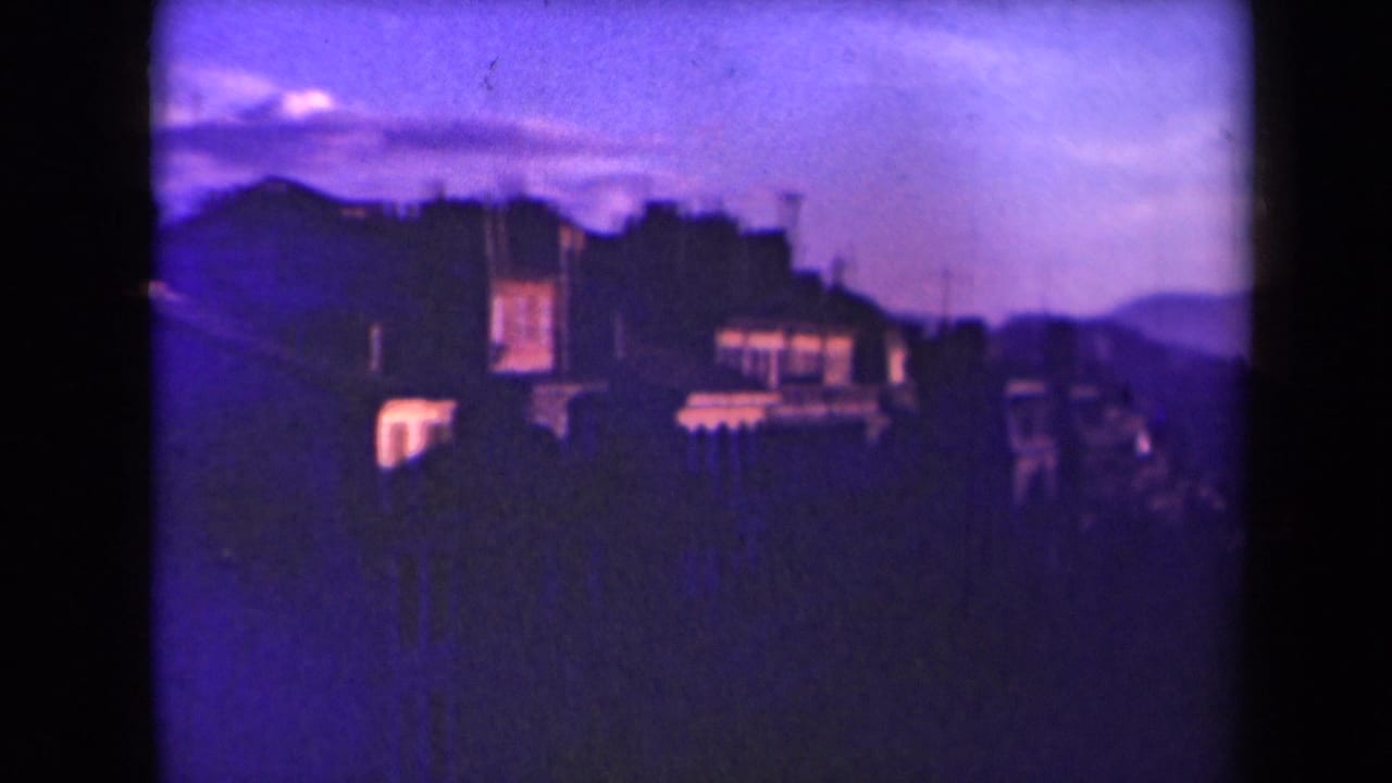 Purple-tinted dusk view of rooftops and distant hills behind dark city silhouettes.