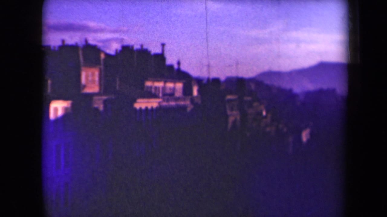 Dusk cityscape with dark rooftops, lit window areas, and hills beyond.