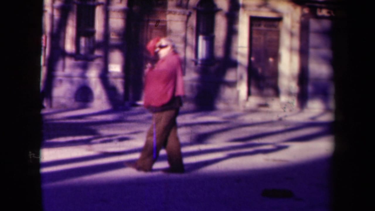 1945 France street frame showing a lone man in a red jacket and dark trousers walking past historic stone facades in long shadows.