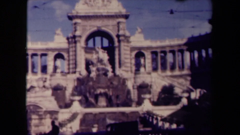 1945 France 8mm frame with a closer street-level view of an ornate stone building, arched window, columns, terrace, and deep shadows.
