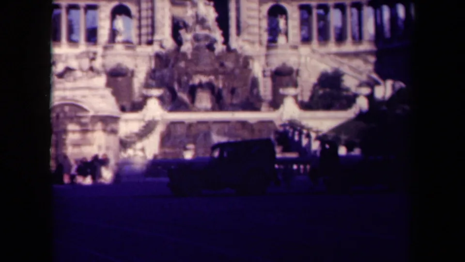 1945 8mm travel frame in France with a sculptural fountain, ornate facade, tiny people, and a dark car crossing the foreground.
