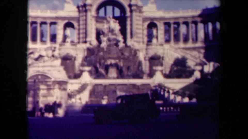 Soft 1945 France home-movie frame showing the fountain and classical facade with a dark car and tiny people in the foreground.