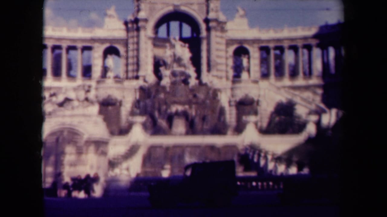 1940s France home-movie frame showing an ornate arched public monument with sculptural fountain elements and street activity below.