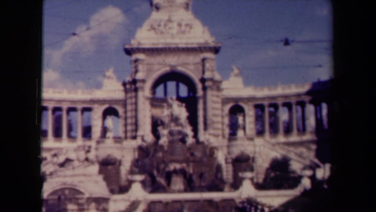 1940s France home-movie frame showing a closer street-level view of the arched monument and central sculptural fountain.