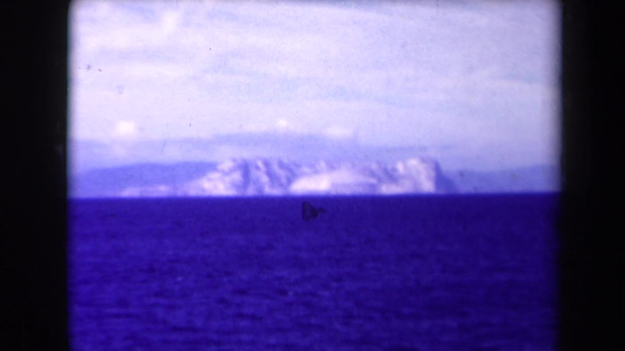Distant view appears to show the Rock of Gibraltar across blue Mediterranean water, filmed from offshore in 8mm color.