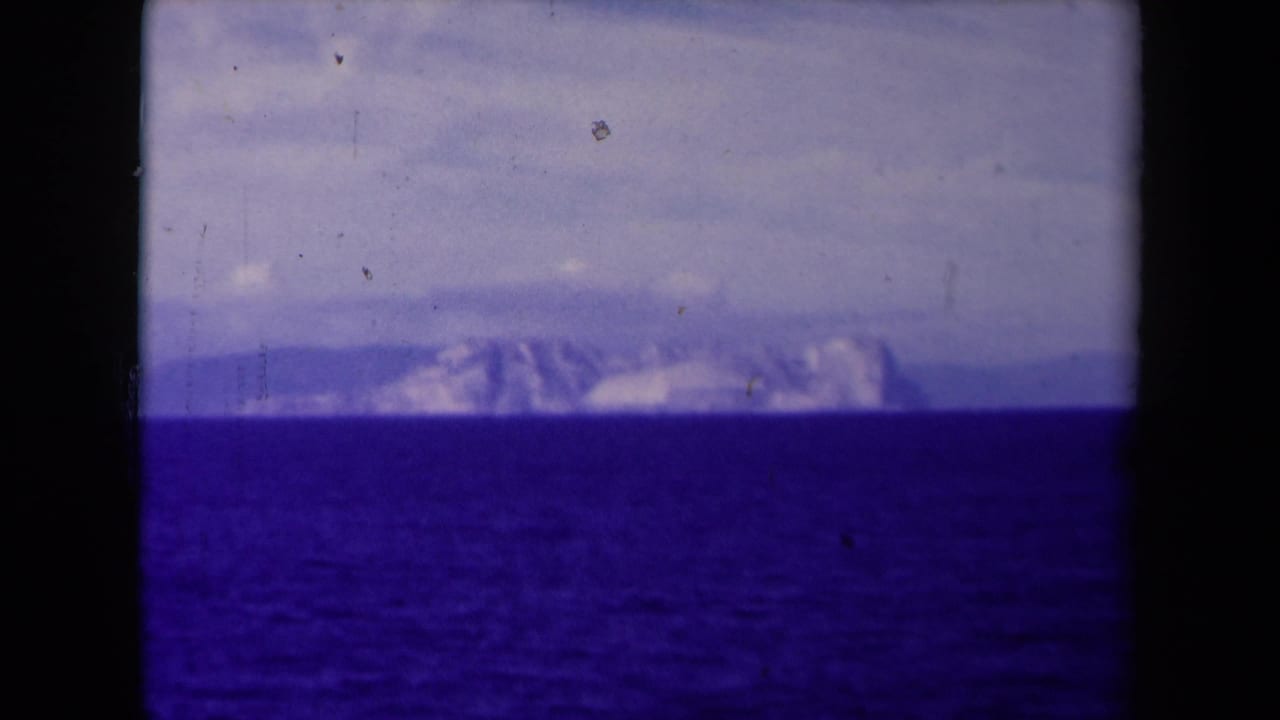 The 1945 8mm frame shows a low, distant mountainous horizon over dark water, softened by haze, grain, and blue-purple film shift.