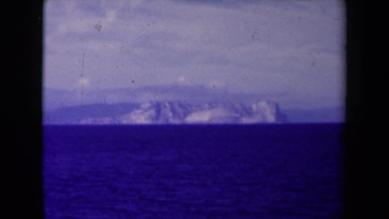 A final 8mm frame holds on rugged distant mountains across the sea, with cloudy sky, film grain, and dark vertical frame edges.